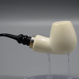May include: A white ceramic pipe with a black mouthpiece and a silver band. The pipe is sitting on a clear stand.
