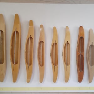 May include: A collection of nine light-colored wooden weaving shuttles arranged in a row, each featuring a central metal rod. A yellow measuring tape is positioned vertically on the left, showing lengths ranging from approximately 10 to 30 centimeters.