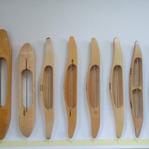 May include: Eight light-colored wooden weaving shuttles of varying lengths and shapes arranged in a row. A yellow measuring tape is positioned vertically on the left, showing the largest shuttle measures approximately 15 inches in length.