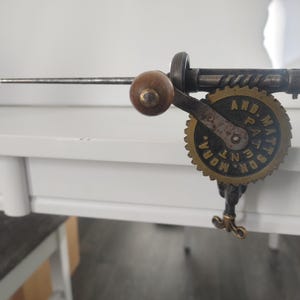 Pode incluir: Uma ferramenta manual de metal vintage com cabo de madeira, uma haste metálica horizontal e uma engrenagem circular com o texto AND. MATTSON, MORA, PATENT. O dispositivo inclui um mecanismo de grampo de parafuso para fixar na borda.
