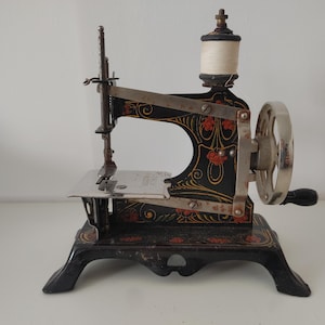 Könnte beinhalten: Antike Nähmaschine in Schwarz und Gold mit floralen Verzierungen. Sie verfügt über eine Handkurbel, eine Garnspule und eine silberne Nadelplatte. Der verzierte Sockel deutet auf das frühe 20. Jahrhundert hin.