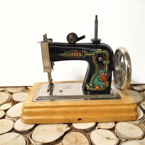 Könnte beinhalten: Eine schwarze Spielzeug-Nähmaschine mit einem Holzsockel. Die Maschine hat eine silberne Nadelplatte und ein silbernes Handrad. Die Maschine ist mit "GLORIA" in goldenen Buchstaben beschriftet.