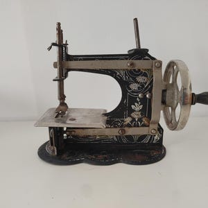 Op de afbeelding: Antieke zwart-zilveren naaimachine met bloemmotieven. De machine heeft een handslinger, een naald en een kleine zilveren basis. Het ontwerp omvat decoratieve bloemmotieven en een zilveren handwiel.