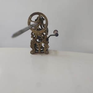 Op de afbeelding: Een vintage mechanisch apparaat van messing met een sierlijk frame, een cirkelvormig tandwiel en een centrale metalen staaf die naar voren uitsteekt. Een kleine handslinger met een donker handvat is aan de zijkant bevestigd.