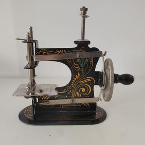 Op de afbeelding: Een miniatuur antieke naaimachine. De zwarte metalen behuizing heeft gouden en groene bloemmotieven. Het heeft een zilveren naald, een handwiel en een kleine basis. Een vintage item voor verzamelaars.