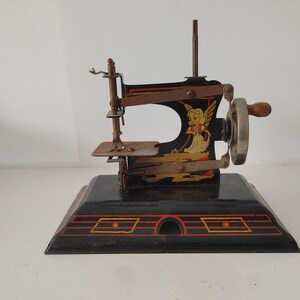 Könnte beinhalten: Antike Spielzeug-Nähmaschine mit schwarzem Sockel und einem dekorativen Design mit einem Engel. Die Maschine hat eine Handkurbel und filigrane Metalldetails, die eine Vintage-Ästhetik zeigen.