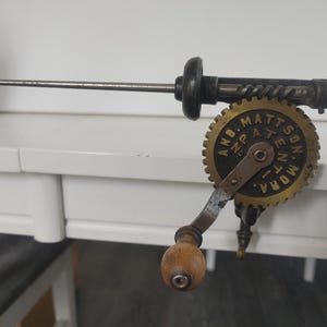Può includere: Un utensile da perforazione manuale vintage in metallo con un'asta in acciaio orizzontale, un corpo in metallo scuro e una ruota dentata color ottone con il testo in rilievo AND. MATTSON PATENT MORA. La manovella ha un pomello in legno chiaro.