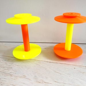 Könnte beinhalten: Zwei spulenförmige Objekte aus glattem Kunststoff. Die linke Spule hat eine leuchtend gelbe Basis und Oberseite mit einer orangefarbenen Mittelsäule. Die rechte Spule hat eine leuchtend orangefarbene Basis und Oberseite mit einer gelben Mittelsäule. Beide Gegenstände bestehen aus einer flachen, kreisförmigen Scheibe oben und unten, die durch einen zylindrischen Mittelpfosten verbunden sind.