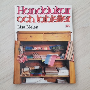 May include: A book titled "Handdukar och tabletter" by Lisa Melén, with an image of a wooden cabinet filled with colorful towels and tablecloths. The book cover features the ICA logo.