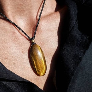 May include: A pendant necklace featuring a polished, oval-shaped tiger's eye stone. The pendant is suspended from a black woven cord. The stone displays golden-brown hues with natural patterns. The necklace is worn against a dark background.