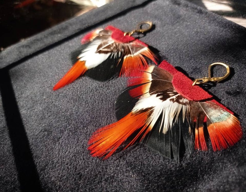 May include: A pair of handcrafted feather earrings. The earrings feature a combination of red, white, black, and brown feathers, with a small red suede accent and gold-colored ear hooks. The feathers are arranged to create a unique, eye-catching design.