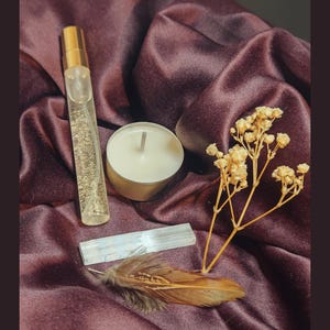 May include: A clear glass perfume atomizer with a gold cap, a white tea light candle in a metal cup, a rectangular selenite crystal wand, a small sprig of dried baby's breath flowers, and a single brown bird feather.