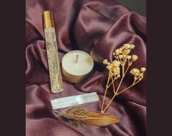May include: A clear glass perfume atomizer with a gold cap, a white tea light candle in a metal cup, a rectangular selenite crystal wand, a small sprig of dried baby's breath flowers, and a single brown bird feather.