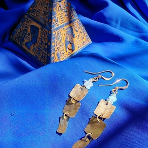 May include: A pair of dangle earrings featuring three graduated brass-toned square metal plates linked together, accented with a small pale blue faceted bead near the gold-toned ear wire hook, showcasing a metallic finish and geometric design.