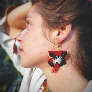 May include: A close-up of a pair of feather earrings. The earrings feature a red, black, white, and teal color scheme. The earrings are attached to a gold-colored hook. The earrings are a unique accessory.