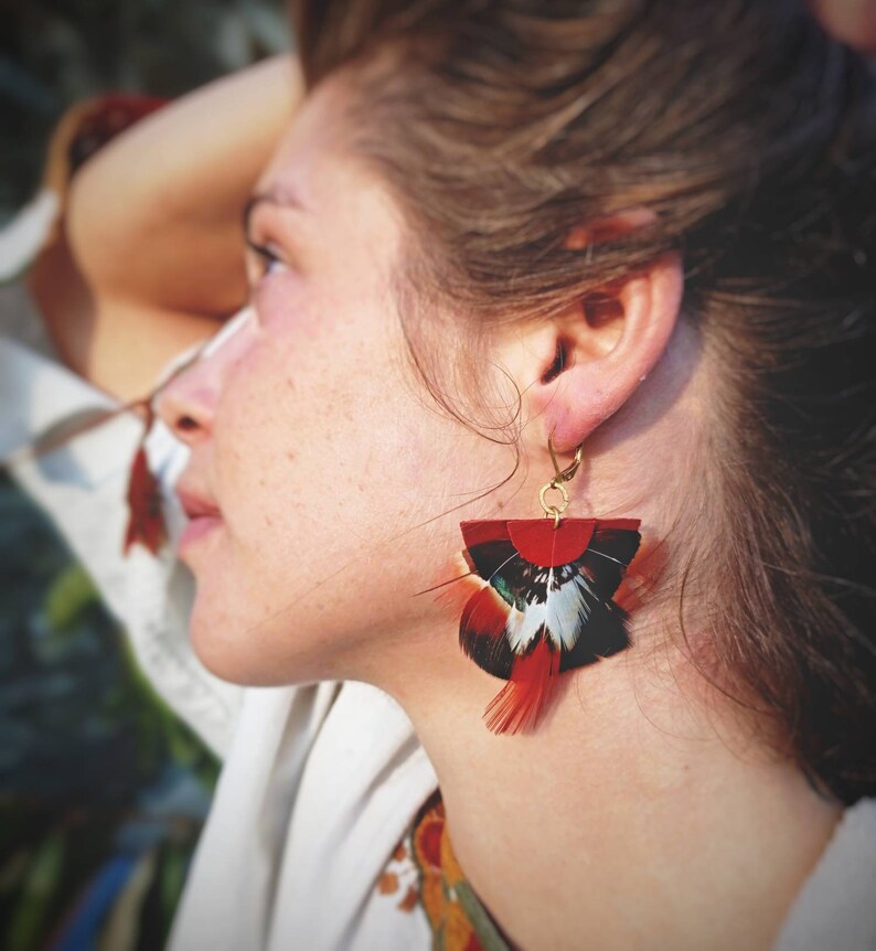May include: A close-up of a pair of feather earrings. The earrings feature a red, black, white, and teal color scheme. The earrings are attached to a gold-colored hook. The earrings are a unique accessory.