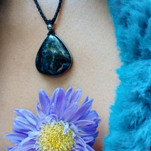 May include: A teardrop-shaped dark blue and black stone pendant suspended from a thin black braided cord. The pendant features a small metallic bead at the top. Below the pendant, a light purple flower with a yellow center is visible.