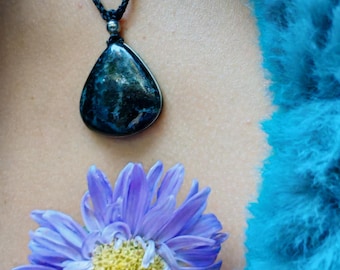 May include: A teardrop-shaped dark blue and black stone pendant suspended from a thin black braided cord. The pendant features a small metallic bead at the top. Below the pendant, a light purple flower with a yellow center is visible.