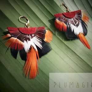 May include: A pair of handmade feather earrings. Each earring features a burgundy suede top, with a fan of black, white, and orange feathers. The earrings have gold-colored lever-back closures. The earrings are displayed on a green leaf.