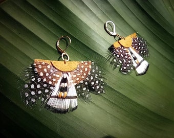 May include: A pair of handmade earrings featuring a fan-shaped design made from feathers. The feathers are primarily white with black spots and brown accents, attached to a tan leather backing with gold-colored ear wires. The earrings are displayed on a green leaf.