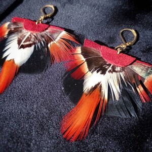 May include: A pair of handmade feather earrings. Each earring features a burgundy suede top with a fan of feathers in black, white, and orange. The earrings have gold-colored ear wires and are displayed on a dark blue surface.