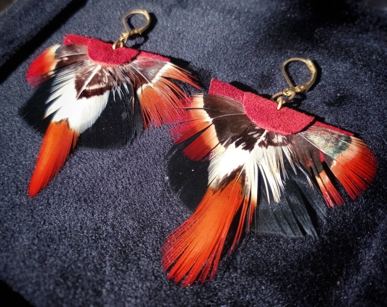 May include: A pair of handmade feather earrings. Each earring features a burgundy suede top with a fan of feathers in black, white, and orange. The earrings have gold-colored ear wires and are displayed on a dark blue surface.