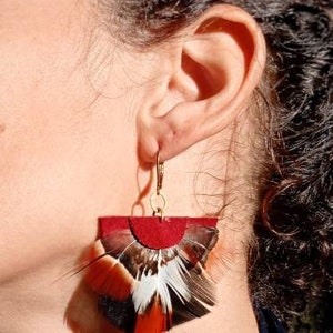 May include: Earrings with a red suede top and a fan of feathers in black, white, and orange. The earrings hang from gold-colored hooks. The earrings are a unique accessory.