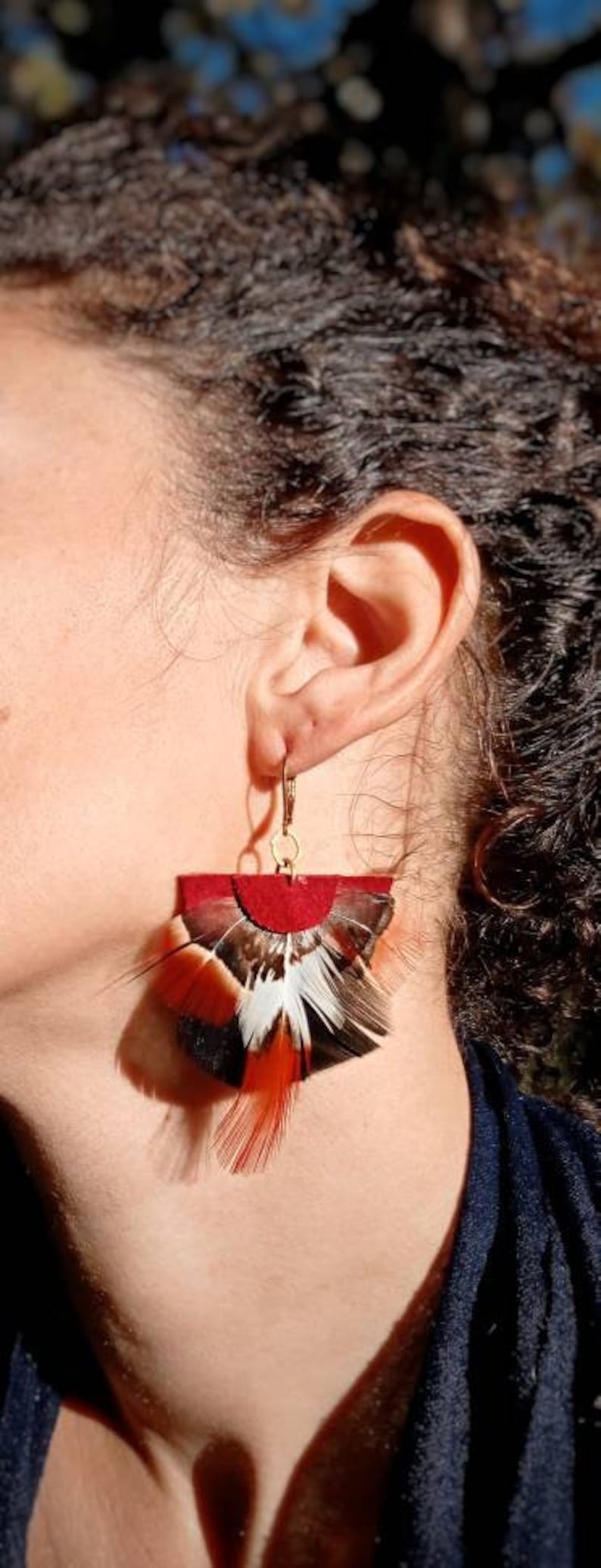 May include: Earrings with a red suede top and a fan of feathers in black, white, and orange. The earrings hang from gold-colored hooks. The earrings are a unique accessory.