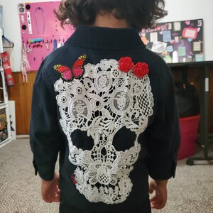 May include: A black button-up shirt with a white lace skull design on the back. The skull has a pink butterfly and red flower embellishments.
