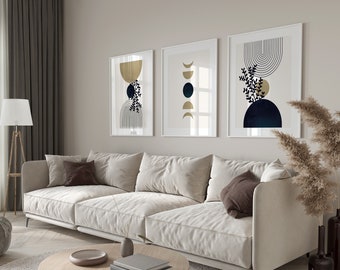 Op de afbeelding: Een witte sectionele bank met bruine kussens staat voor een muur met drie ingelijste abstracte kunstprints met gouden en blauwe geometrische vormen. Rechts van de bank staat een gedroogde pampasgras plant in een vaas.