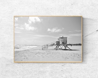 May include: A black and white photograph of a lifeguard stand on a sandy beach. The stand is a simple structure with a roof and a ladder leading up to the top. The beach is empty except for the stand and some footprints in the sand.