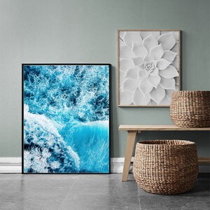 May include: Two framed prints, one with a blue and white abstract ocean wave design and the other with a white flower design. The prints are hanging on a light gray wall above a wooden shelf with two woven baskets.