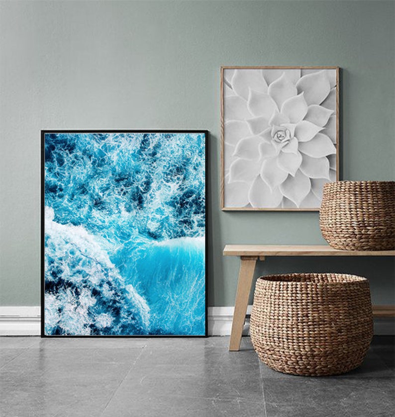 May include: Two framed prints, one with a blue and white abstract ocean wave design and the other with a white flower design. The prints are hanging on a light gray wall above a wooden shelf with two woven baskets.