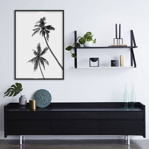 May include: Black and white framed print of two palm trees against a white background. The print hangs above a black cabinet with a marble clock, a gold cylinder, and a gray vase. A black shelf with plants and candles is also visible.