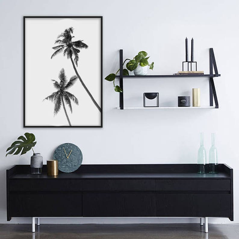 May include: Black and white framed print of two palm trees against a white background. The print hangs above a black cabinet with a marble clock, a gold cylinder, and a gray vase. A black shelf with plants and candles is also visible.