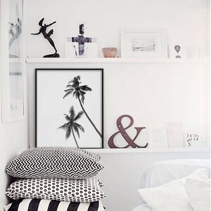 May include: A black and white framed print of two palm trees, displayed on a white wall. Below the print are three decorative pillows with geometric patterns. A shelf above holds various decorative items.