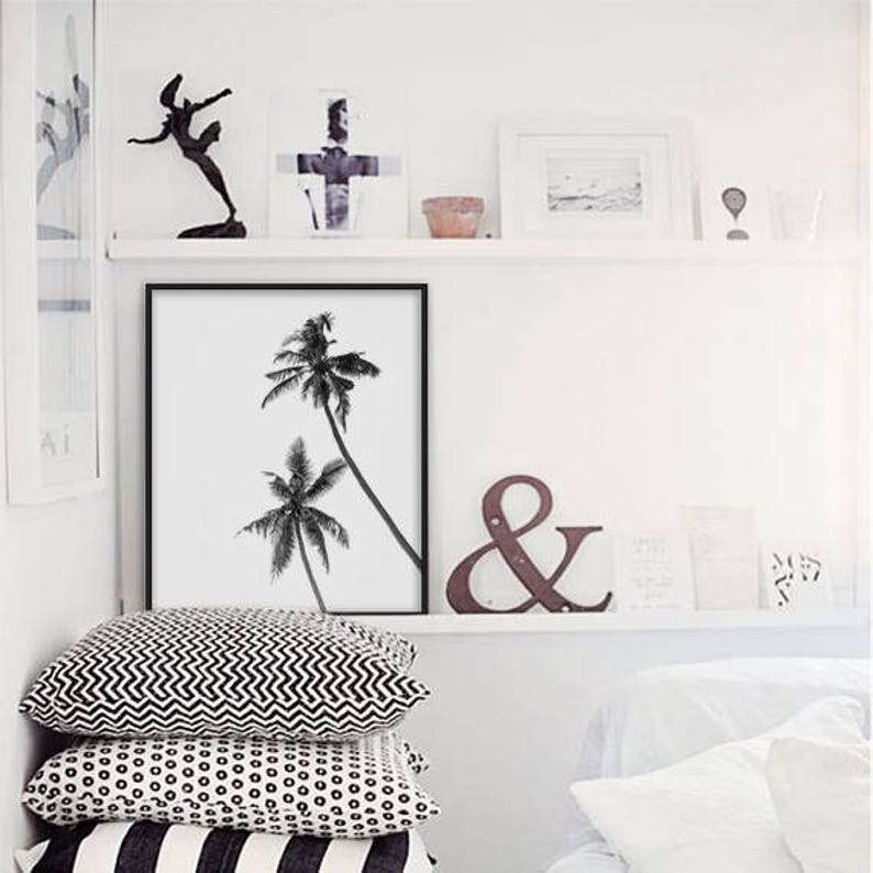 May include: A black and white framed print of two palm trees, displayed on a white wall. Below the print are three decorative pillows with geometric patterns. A shelf above holds various decorative items.