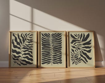 Op de afbeelding: Een set van drie rechthoekige wandkunstprints in lichte houten lijsten. Elke print toont een botanische illustratie van donker houtskoolzwarte bladeren tegen een effen lichtbeige achtergrond met duidelijke varenachtige patronen.
