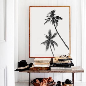 May include: A framed black and white print of two palm trees leans against a white wall. A rustic wooden bench holds a stack of books, a black hat, a woven bag, a wooden bowl with fabric, and black sandals.