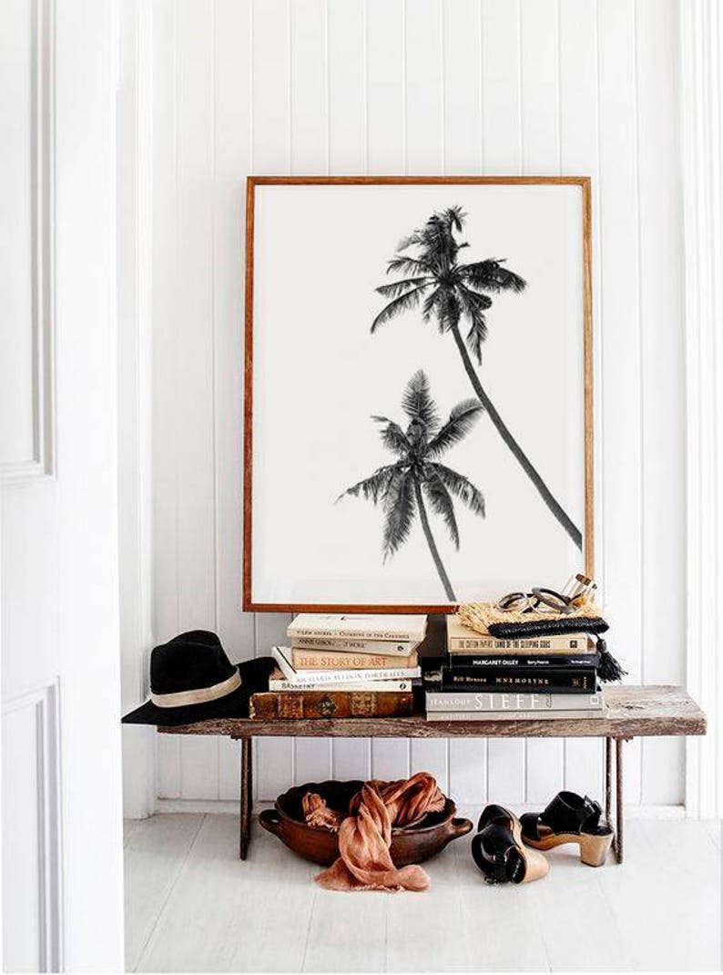 May include: A framed black and white print of two palm trees leans against a white wall. A rustic wooden bench holds a stack of books, a black hat, a woven bag, a wooden bowl with fabric, and black sandals.