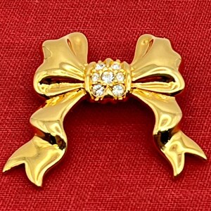 Op de afbeelding: Een gouden strik broche met een cluster van heldere strass steentjes in het midden.