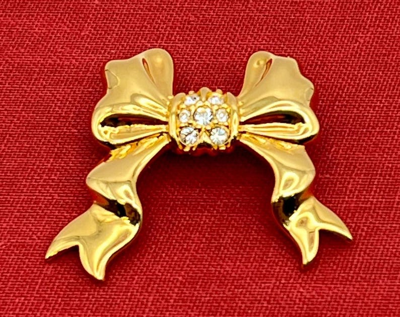 Op de afbeelding: Een gouden strik broche met een cluster van heldere strass steentjes in het midden.