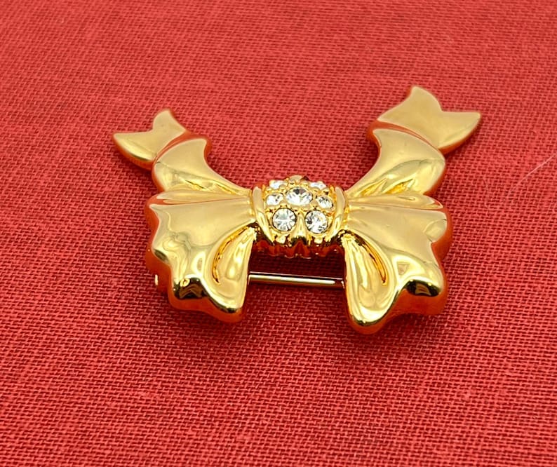Op de afbeelding: Een gouden strik broche met een helder strass steentje in het midden.