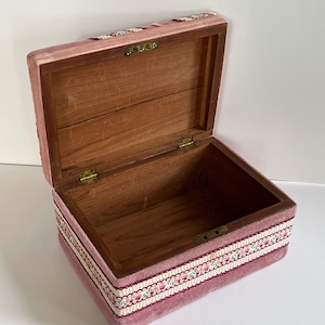 Peut inclure: Une boîte à bijoux en velours rose avec une bordure florale et un intérieur en bois. La boîte est ouverte et le couvercle est relevé.