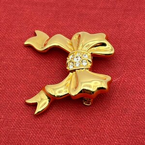 Op de afbeelding: Een gouden strik broche met heldere strass steentjes in het midden. De strik is gemaakt van glad, glanzend metaal.