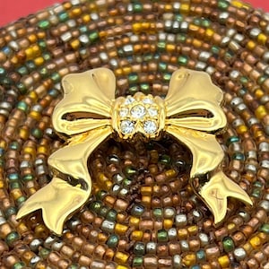 Op de afbeelding: Een gouden strik broche met een helder strass steentje in het midden. De broche is geplaatst tegen een achtergrond van bruine en groene kralen.
