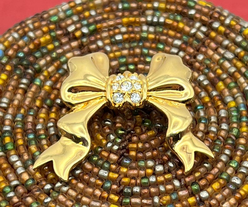 Op de afbeelding: Een gouden strik broche met een helder strass steentje in het midden. De broche is geplaatst tegen een achtergrond van bruine en groene kralen.