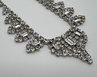May include: A silver-toned necklace with a delicate chain and a series of sparkling, clear rhinestones. The necklace features a central design with larger, rectangular stones surrounded by smaller, round stones, creating a glamorous look.