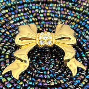 Op de afbeelding: Een gouden strik broche met een helder strass steentje in het midden. De broche is geplaatst op een achtergrond van iriserende kralen.