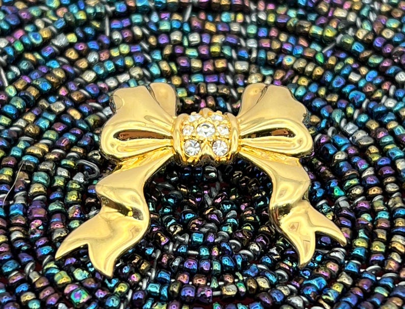 Op de afbeelding: Een gouden strik broche met een helder strass steentje in het midden. De broche is geplaatst op een achtergrond van iriserende kralen.
